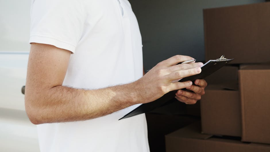 A person in a white short-sleeved shirt holds a black clipboard and pen, standing in an indoor residential setting with a background of stacked brown cardboard moving boxes and packaging material. The individual appears to be inspecting or making notes related to a home relocation or furniture transport process, capturing the typical scene of packing and moving activities associated with house removals. The environment is well-lit, with visible details of the boxes arranged on the floor, indicating an ongoing packing or inventory check as part of a professional removals service. This scene illustrates the planning and organisational stage of a home relocation, often managed by companies like Fulham Man and Van, which specialise in removals and moving logistics, including packing, loading, and transport coordination.