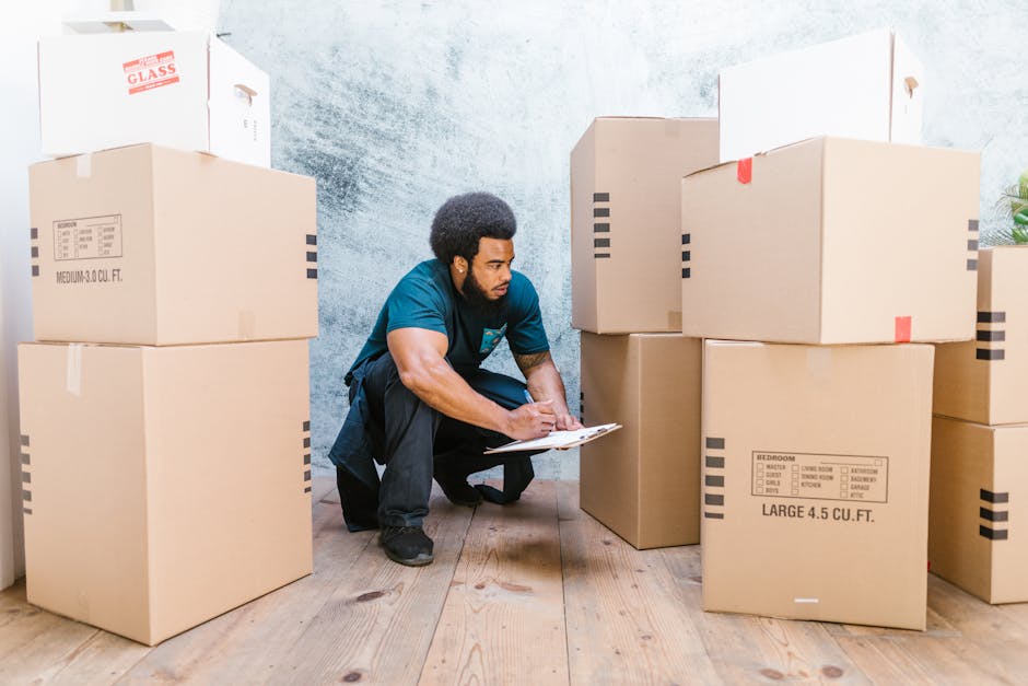 A person in a white short-sleeved shirt holds a black clipboard and pen, standing in an indoor residential setting with a background of stacked brown cardboard moving boxes and packaging material. The individual appears to be inspecting or making notes related to a home relocation or furniture transport process, capturing the typical scene of packing and moving activities associated with house removals. The environment is well-lit, with visible details of the boxes arranged on the floor, indicating an ongoing packing or inventory check as part of a professional removals service. This scene illustrates the planning and organisational stage of a home relocation, often managed by companies like Fulham Man and Van, which specialise in removals and moving logistics, including packing, loading, and transport coordination.