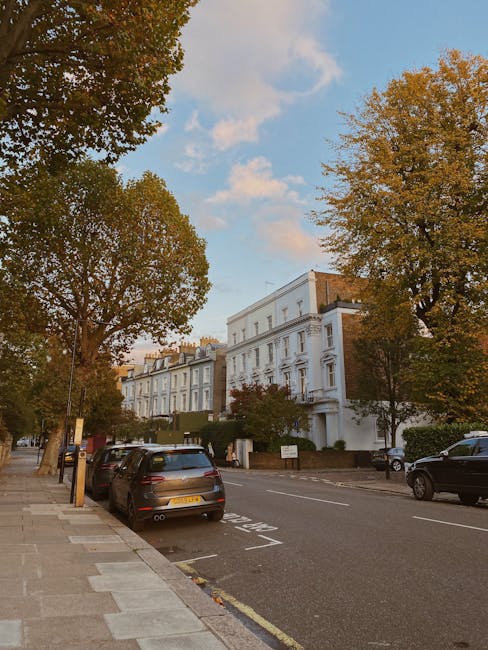 A street scene in Fulham with a row of white residential buildings, some with decorative architectural details, lined with large trees displaying autumn foliage in shades of green, yellow, and orange. Parked cars, including a dark-colored hatchback and a black SUV, are along the curb on a paved sidewalk. The sky overhead is partly cloudy with patches of blue visible. In the context of home relocation and moving services, Fulham Man and Van is involved in the transportation and loading process, likely preparing to load furniture and boxes into a van parked nearby, supported by moving blankets and equipment visible on site.