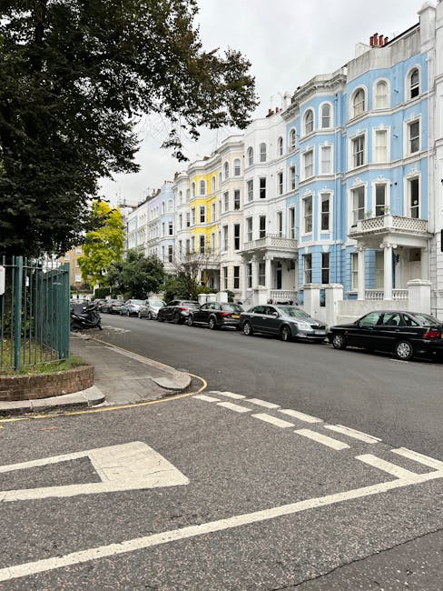 A quiet residential street with a row of traditional brick and stone terraced houses on the left, each with small front gardens filled with green shrubs and flowering plants. A tall, slightly weathered lamppost stands near the curb, casting shadows on the smooth tarmac road. The street is lined with lush, mature trees on both sides, with vibrant green leaves providing shade and creating a canopy effect. In the background, additional buildings are visible along the street, with the clear blue sky overhead indicating a bright, sunny day. The environment suggests a peaceful, suburban neighbourhood suitable for home relocation and furniture transport services. Complete with natural lighting, the scene reflects an ideal setting where house removals, packing, and loading operations by companies like Fulham Man and Van could take place, involving careful handling of belongings within this scenic area.