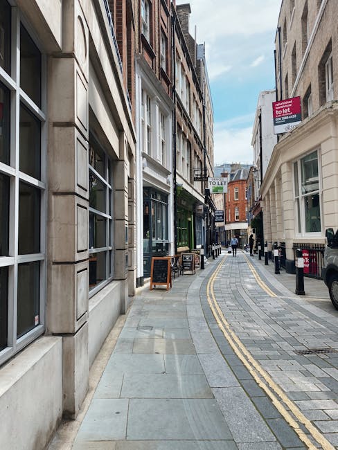 A narrow street in Fulham with a curved sidewalk paved with grey and beige bricks, lined by multi-storey buildings constructed from brick and stone, featuring large windows and decorative facades. On 