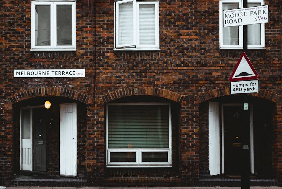 A brick townhouse on Melbourne Terrace SW6 showing two white-framed windows on the upper floor, with the lower level featuring a white door on the left and a large window with closed blinds in the center. An exterior wall-mounted light is situated next to the door, illuminating the entrance area. Two street signs are visible: one indicating Melbourne Terrace SW6 and another for Moore Park Road SW6, which is mounted higher up and slightly to the right. A traffic warning sign below the Moore Park Road sign displays a speed bump with the text 'Humps for 450 yards.' The building's façade is composed of dark red and brown bricks, and the scene appears to be captured during dusk or low-light conditions. The setting suggests an urban residential area where home relocation or furniture transport could be taking place, typical of house removals or moving services in Fulham, as operated by Fulham Man and Van.