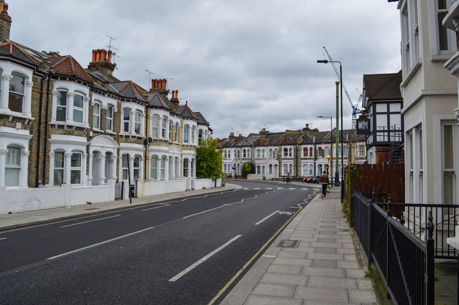 A quiet residential street in Fulham featuring Victorian-style terraced houses with bay windows and brick facades, some with white-painted trim and decorative architectural details. The street is lined with a grey pavement on the right, separated from the road by a black wrought iron fence. There are a few pedestrian figures walking along the pavement, and parked cars are visibleFurther down the street. The sky overhead is overcast with grey clouds, and street lamps and a construction crane can be seen in the background, indicating ongoing building work. This setting provides a typical scene for house removals or relocation services, with the quiet street illustrating the environment where Fulham Man and Van might operate for home relocation and furniture transport tasks.