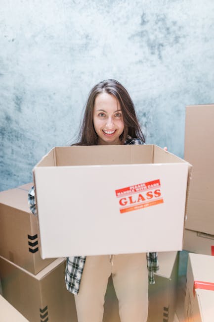 A smiling woman with long brown hair stands indoors, holding a large cardboard box labeled 'Fragile: Glass' in red text. She is wearing a checkered shirt over a white top and light-colored trousers. Behind her, there are several other packed cardboard boxes of various sizes stacked on the floor, some with black tape securing the lids. The background features a textured, light blue wall, indicating an interior space prepared for a home relocation. The scene captures the process of packing and moving, with the woman actively involved in furniture transport or packing tips, as part of a house removals service by Fulham Man and Van, illustrating the logistics of home removals and packing during a house move.