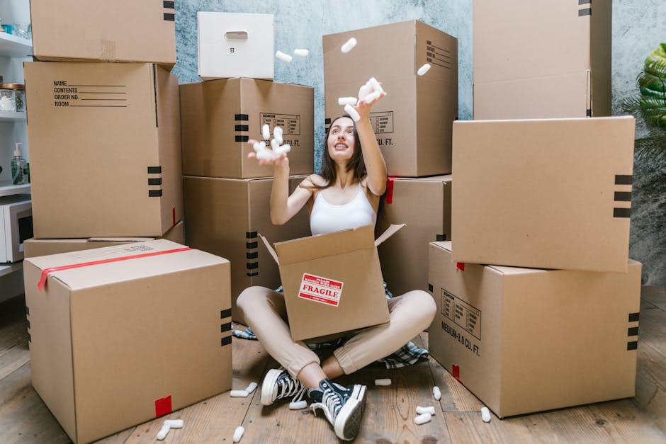 A young woman sits on a wooden floor inside a home surrounded by numerous cardboard moving boxes, some sealed with red tape and others open. She is wearing casual clothes, including a white tank top, beige pants, and sneakers. The woman is smiling while tossing packing foam peanuts into the air, indicating a packing or unpacking process during a furniture removal or home relocation. Behind her, a partially visible doorway leads to another room with a light blue wall, and a small potted plant can be seen on the right side of the image. A red 'Fragile' label is visible on the open box resting on her lap, emphasizing careful handling during furniture transport. This scene illustrates the packing phase of a house move, highlighting the use of cardboard boxes, foam packing materials, and the indoor environment where Fulham Man and Van offers removals and packing services, aligned with the goals of furniture transport and home relocation logistics.