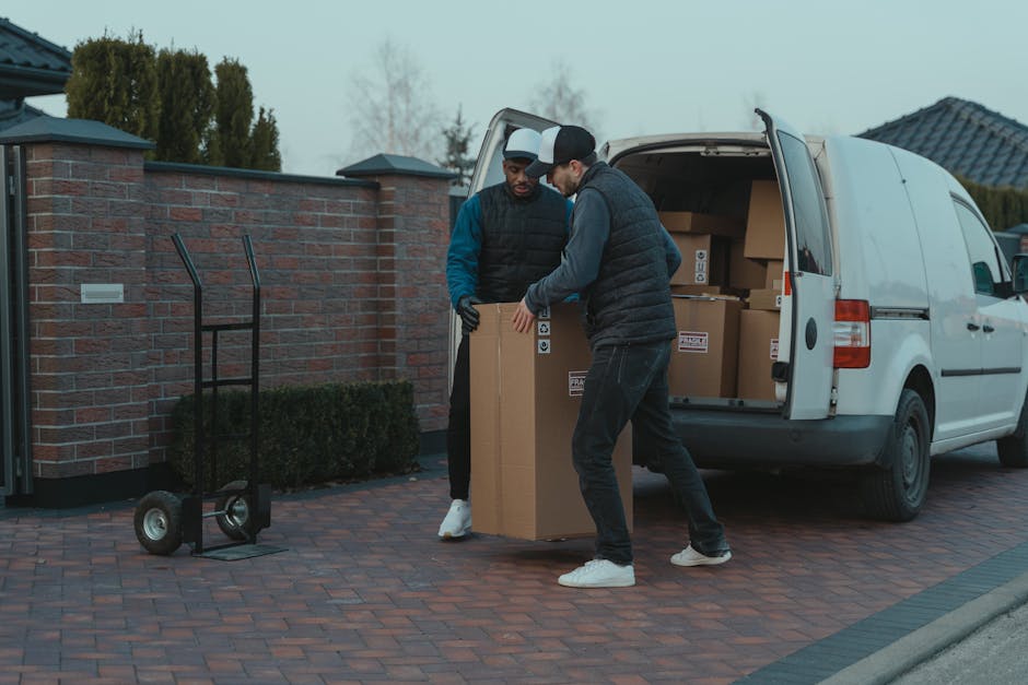 Two men in casual clothing are engaged in a home relocation task outside a residential property, transferring a large cardboard box onto a moving dolly. They are positioned on a brick-paved driveway next to an open white van filled with similarly packed boxes. One man is wearing a black padded vest over a blue jacket and black gloves, while the other is dressed in a grey hoodie with a black quilted vest. The van is parked near a brick wall with a hedge and a house with a dark tiled roof visible in the background under overcast skies. A second empty dolly stands nearby on the pavement, ready for use, with additional boxes stacked inside the van, indicating a packing and moving process managed by Fulham Man and Van. The scene captures the loading process of furniture transport as part of a house relocation, with a focus on careful handling, packaging materials like cardboard boxes, and the vehicle used for house removals.