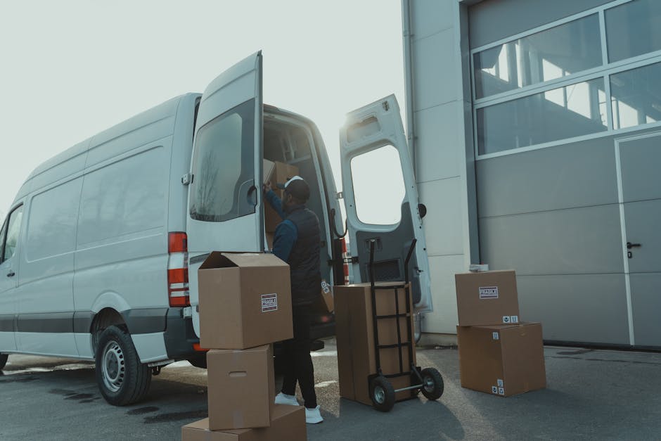A worker loading a white cargo van with cardboard boxes outside a modern building with large glass windows. The worker, dressed in dark clothing with a cap, is handling a box on a black hand truck with a metal frame. Several packed boxes are positioned on the pavement adjacent to the van, some stacked and others awaiting transport. The back doors of the van are wide open, revealing a partially visible interior filled with more moving supplies and packed boxes. The scene is illuminated by natural daylight, with the sun shining from behind the van, casting light onto the loading area. This setup depicts a home relocation process involving furniture transport and packing for a house move, typical of services provided by Fulham Man and Van as part of professional removals or furniture removals and moving logistics.